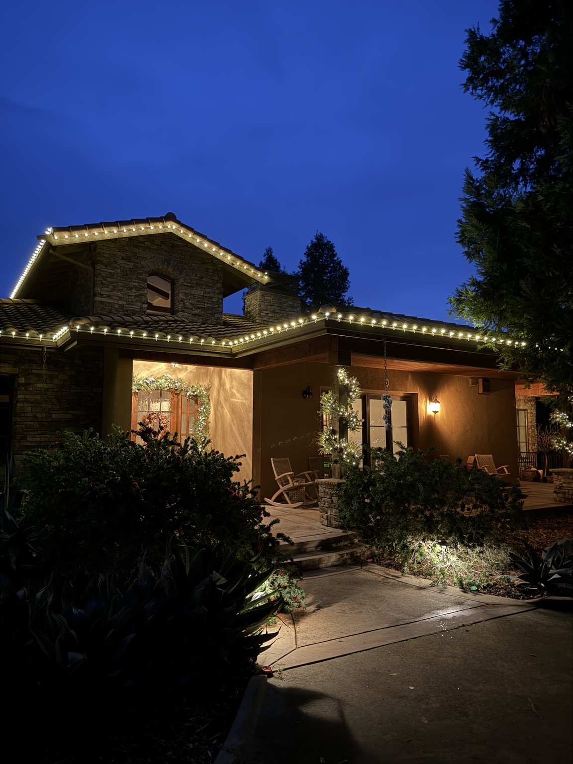 Tile-roof home with warm white C9 Christmas roofline lights, garland and wreaths at dusk