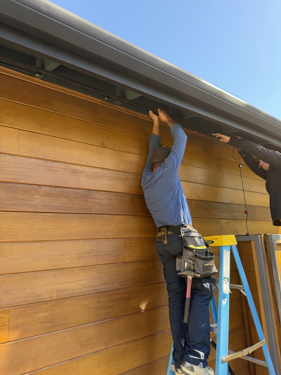 Give It Light installers mounting Govee permanent lighting under a modern wood-clad eave in daylight